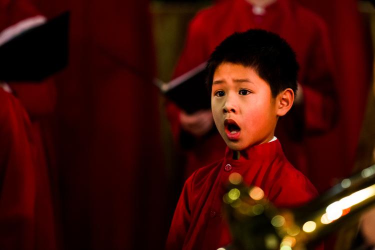 Young boy chorister singing in red robe, with brass instrument in foreground